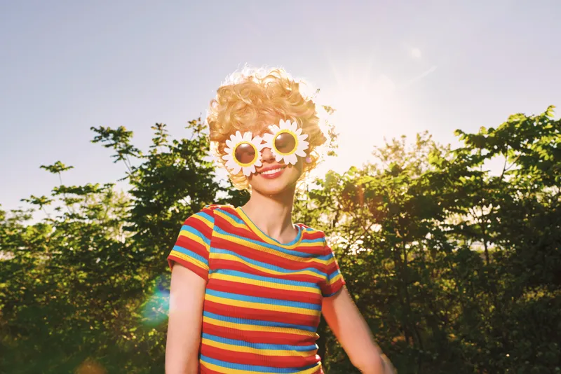 A young woman with blonde curly hair wears a colorful striped shirt and flower sunglasses, standing outdoors in bright sun with lush trees behind and a playful, carefree vibe.