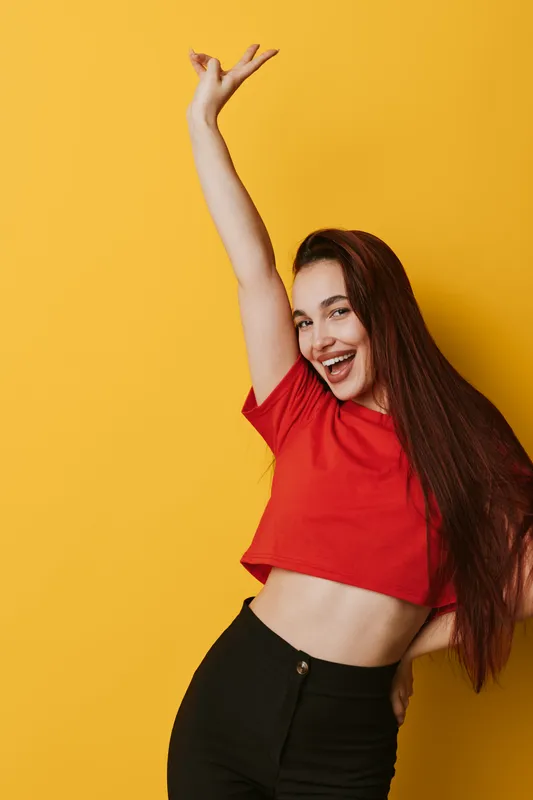Young woman with long hair wearing red crop top and black pants posing cheerfully with raised arm smiling against yellow background in studio. Happy girl expressing joy and energy.