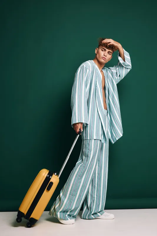 Young man in striped pajamas holding yellow suitcase standing against green background, casual travel outfit, relaxed pose and thoughtful expression in studio setting.