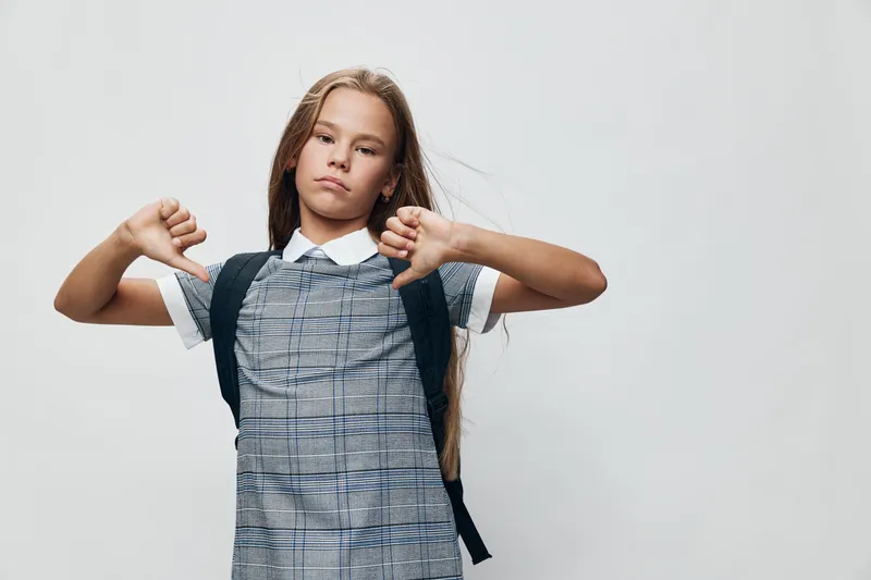 Girl with long hair and school uniform showing thumbs down gesture with both hands wearing backpack on neutral background. Child expressing dislike or disapproval with serious face.