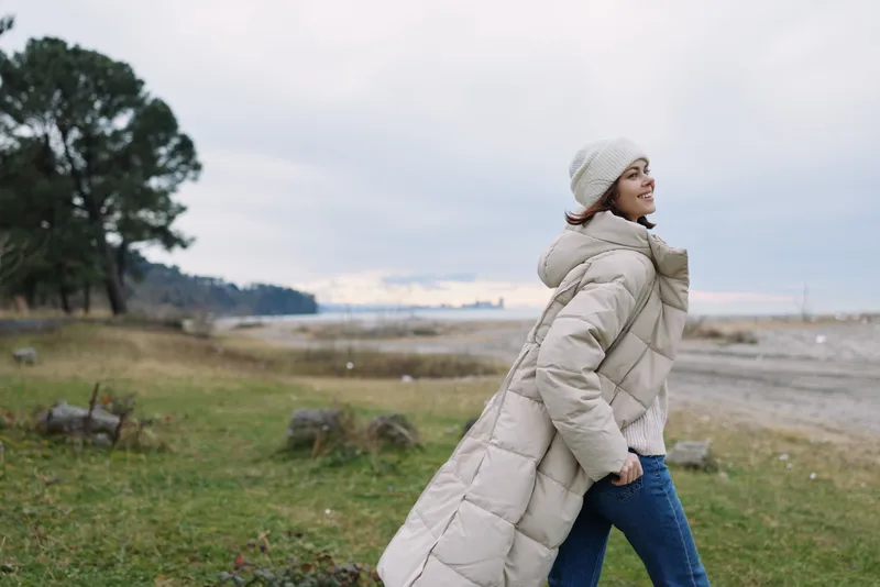 Woman wearing a long beige coat and knit hat walks along a coastal landscape, breezy outdoor scene featuring winter fashion and a calm sea in the background