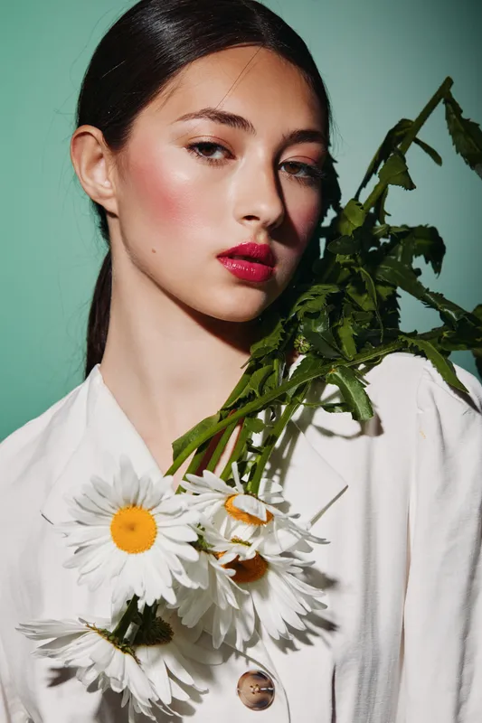 Young woman in white with vivid red lips and flushed cheeks holding fresh daisies. Natural. Artistic beauty concept. Studio shot on green background blending fashion and botany.