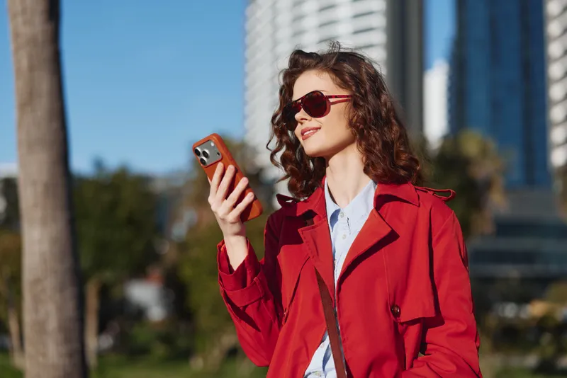 Young woman with curly hair wearing a red coat and sunglasses smiles while using a smartphone outdoors on a sunny day in the city.