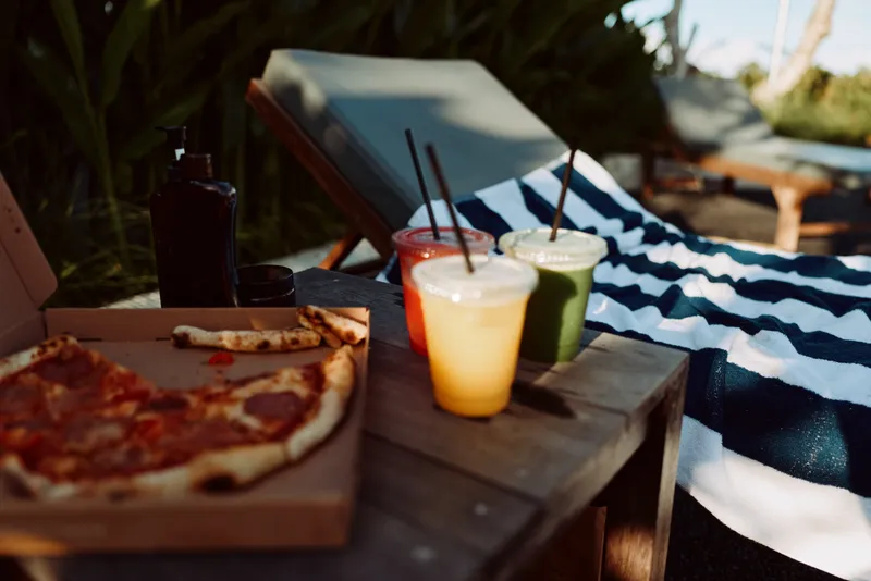 Casual outdoor poolside scene with pizza on a wooden board and colorful drinks, a relaxed friends gathering by lounge chairs, capturing summer leisure and warm mood under sunny skies