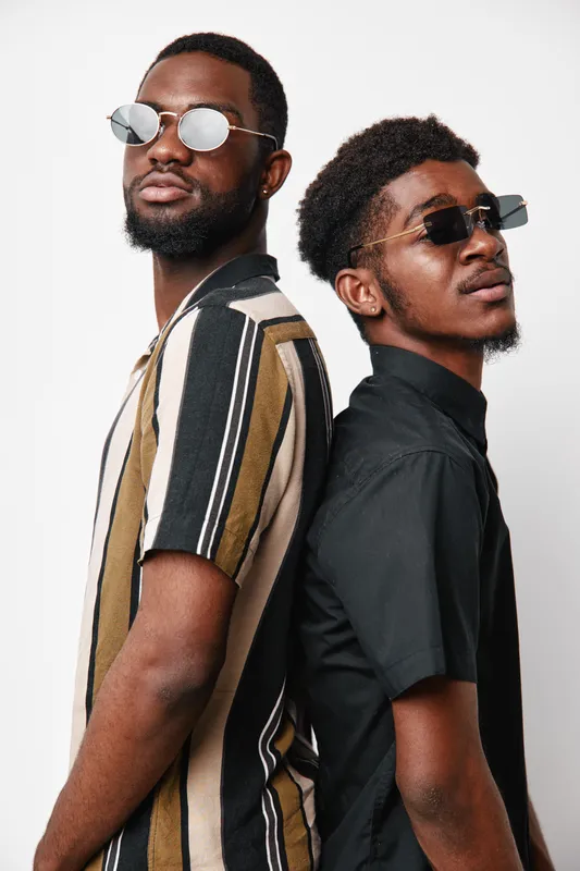 Two young black men with dark skin wearing sunglasses and casual shirts standing back to back against white background. Stylish, confident, and modern male models posing together.