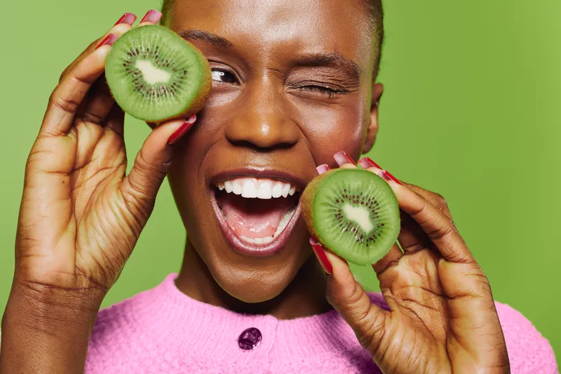 Smiling African woman with dark skin holds two kiwi halves near face, winking, wearing pink sweater on green background. Concept of healthy lifestyle, fresh fruit, and natural beauty.