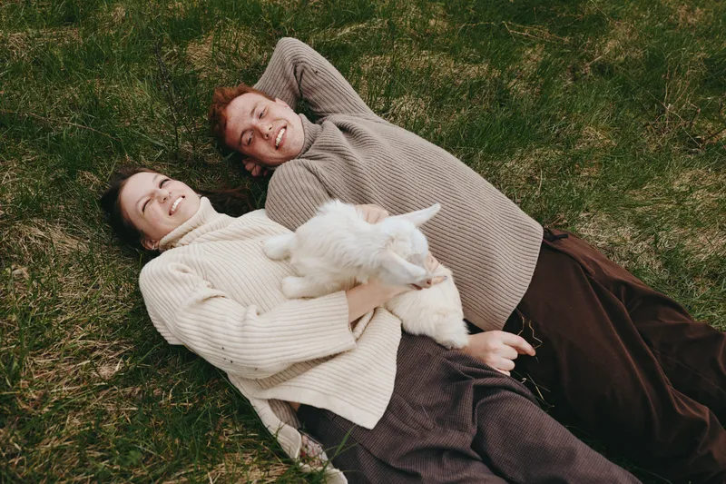 Happy couple relaxing on grass with a playful goat kid, enjoying a cozy outdoor moment, soft sweaters, gentle smiles, and affection, lifestyle scene outdoors. Bright sky above adds mood.