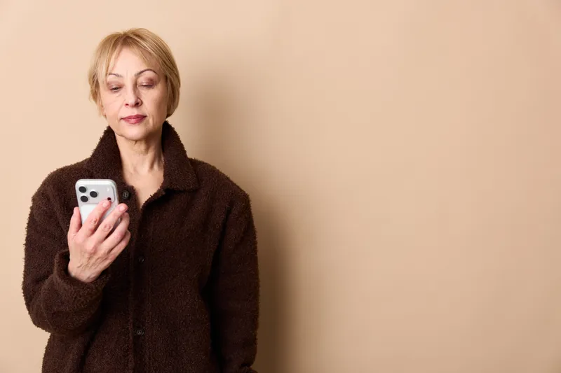 Elderly woman in brown coat holds a smartphone, expression worried and tired, indoor portrait against plain beige background, concept for anxiety, loneliness, communication, and technology