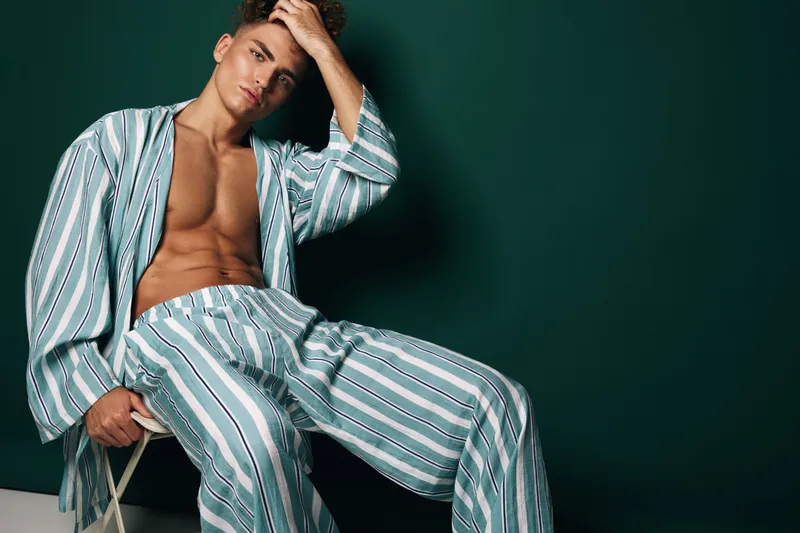 Young man with bare chest wearing striped pajamas sitting on chair against dark background, posing with hand on head in relaxed fashion portrait.