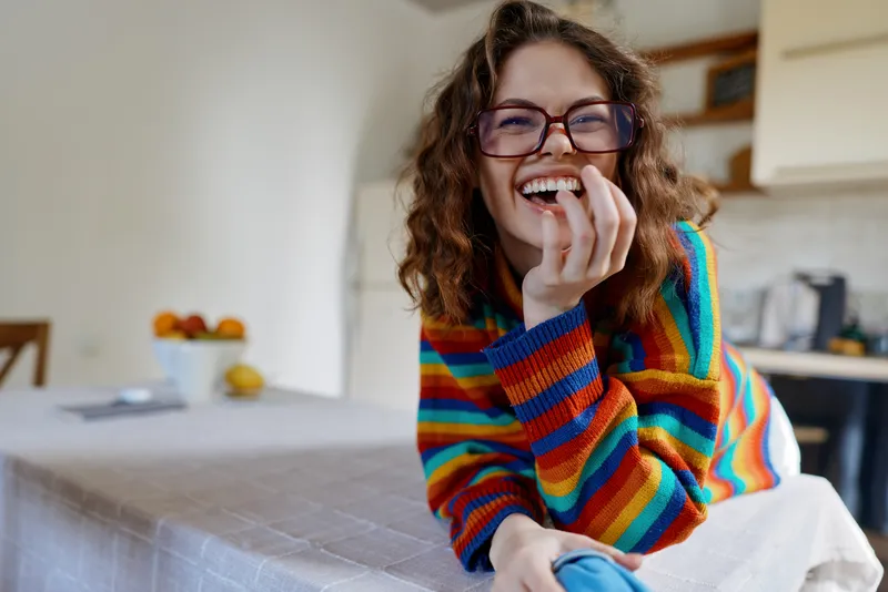 Young woman with curly hair sits at a kitchen table indoors, smiling and relaxed as she rests her hand on her chin