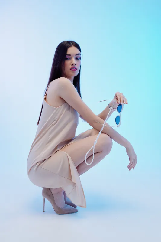 Young woman in a beige dress squats on a blue background holding sunglasses. She wears high heels and has long dark hair. Concept of beauty, fashion, and elegance in studio lighting.