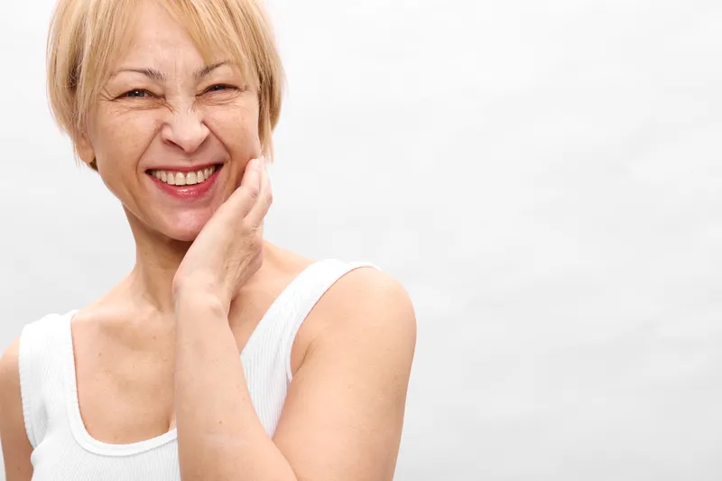 Elderly woman smiling, hand on cheek, gentle skincare concept, wellness and relaxation, bright studio portrait on white background, natural expression, soft copy space