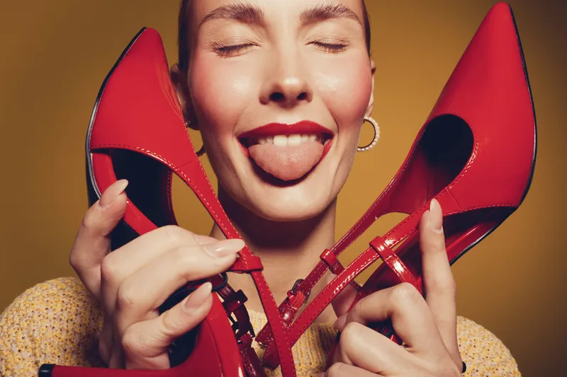 Young woman with closed eyes holds red high heels near face and sticks out tongue. Playful mood with bright yellow background and casual outfit. Concept of fun and fashion.