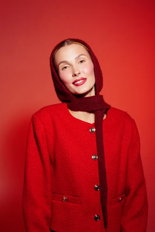 Young woman in red woolen coat and matching scarf poses against red background. She smiles softly with natural makeup and relaxed expression. Concept of fashion and winter style.