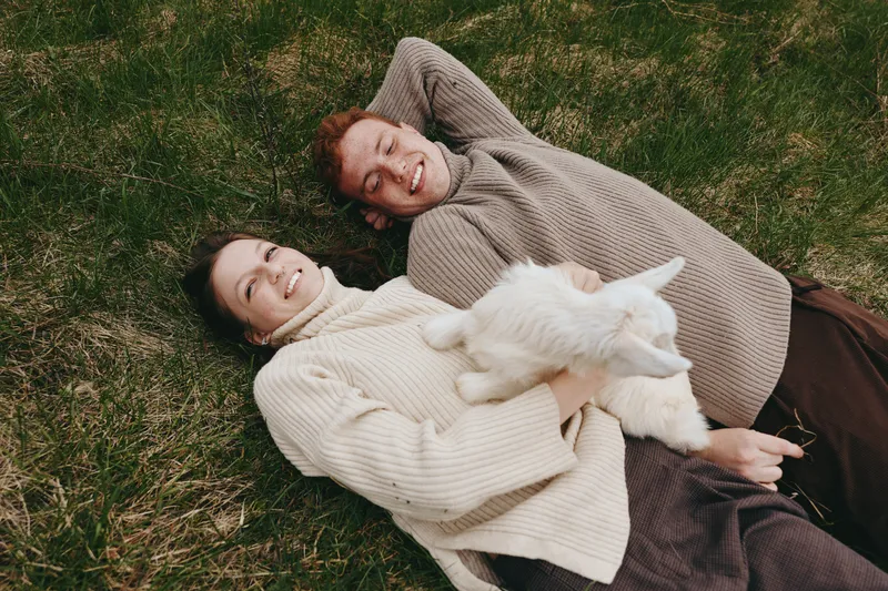 A young woman and man lie on grass smiling while the woman holds a white baby goat, capturing a joyful moment of connection and relaxation in a casual outdoor lifestyle setting.