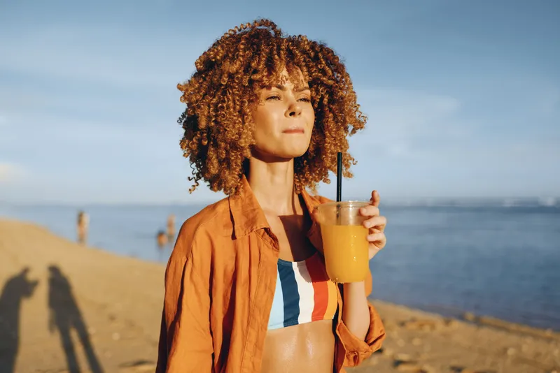 Woman with curly hair holding a mug of hot drink, seaside beach in sunlight, warm jacket, calm relaxed pose, gentle breeze, travel mood, natural colors, coastal horizon, blue sky today