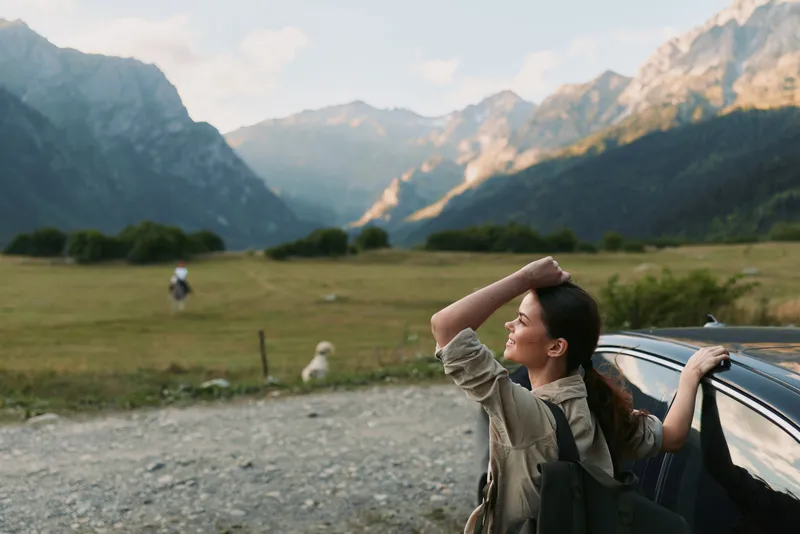 A woman by a car in a mountain valley enjoys the view, embracing a casual outdoors lifestyle and escape into nature, a peaceful moment of exploration and calm.