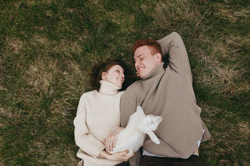 Cheerful couple outdoor lifestyle scene with goat kid, affection and relaxation on grass, greenery and meadow under breeze, warm light, natural tones, candid moment, shared joy, wholesome lifestyle
