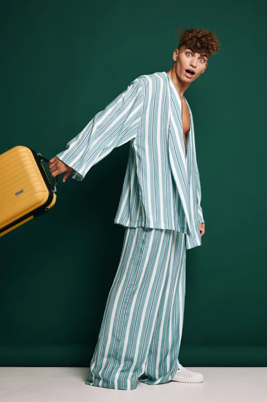 Young man in striped loose suit holding yellow suitcase looking surprised against green background. Fashionable traveler in stylish outfit walking with luggage on barefoot floor.