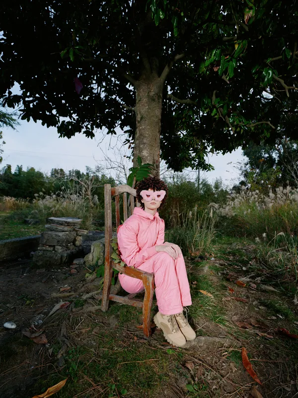 Outdoor portrait of a person in a pink jumpsuit sitting on a wooden chair under a tree, calm countryside setting, casual pose and natural light.