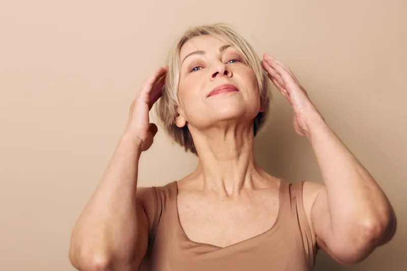 Confident mature woman with short blonde hair touches her head with hands on beige background. Concept of self-care, aging gracefully, beauty, and natural skin confidence.