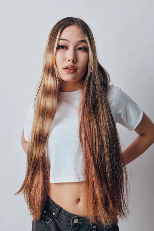 Young woman with long straight hair and casual white t shirt posing indoors on light background with hands on hips looking at camera with relaxed expression and style.