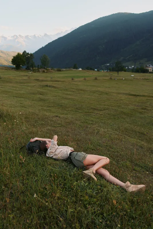 Lying woman in a sunlit meadow, relaxed pose in a wide field with distant mountains, celebrating outdoor lifestyle, natural colors and peaceful countryside under blue sky warm breeze vivid greens.