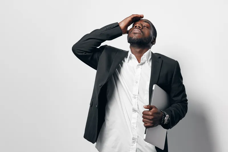 African American man in business suit holding laptop with hand on forehead expressing stress or frustration isolated on white background. Professional black male in formal wear.