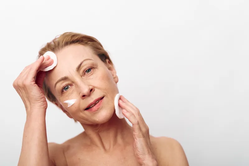 Mature woman with light skin gently cleanses face using cotton pads and cream. Soft natural skincare routine with focus on facial cleansing and self-care on plain white background.