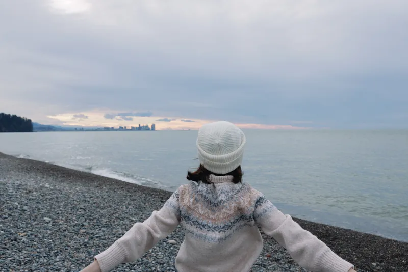 Woman standing on a pebble beach with arms wide, facing the sea and distant skyline under a cloudy sky, wearing a cozy knit sweater and beanie for a peaceful outdoor moment