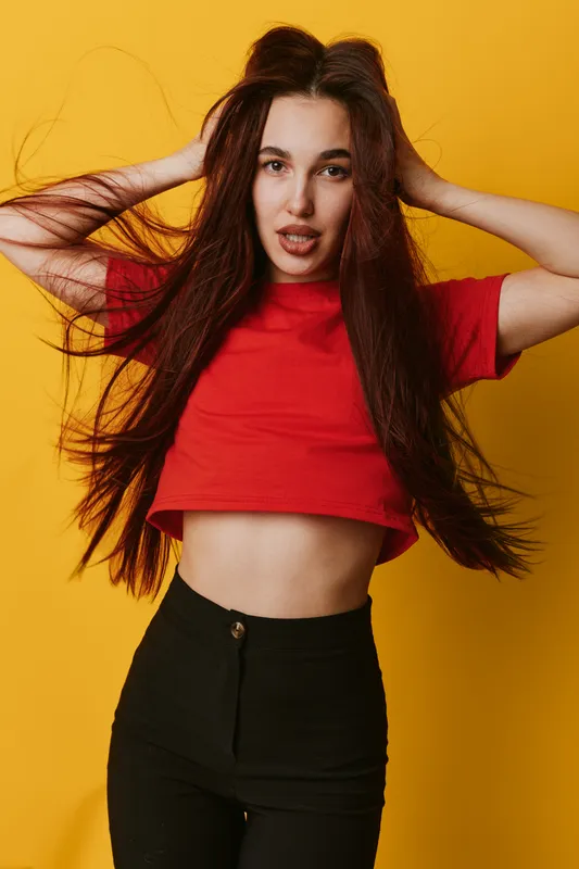 Young woman with long hair wearing red crop top and black pants posing with hands in hair against yellow background for fashion and beauty portrait.
