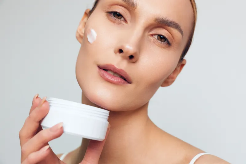 Smiling woman applies white face cream from a jar. Close-up on a neutral background. Skincare concept, self-care, and wellness. Soft studio lighting creates a calm, warm mood and healthy skin.