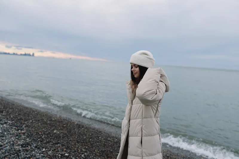 woman, winter, coat, beach, sea, cold, portrait, outdoor, Woman wearing a white puffer coat and knit hat stands on a pebbled shore, watching gentle waves under a cloudy sky at dusk