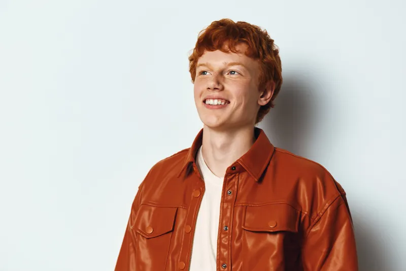 Young man with red hair wearing brown leather jacket smiling and looking right against plain white background in studio. Casual fashion portrait with natural light and positive expression.
