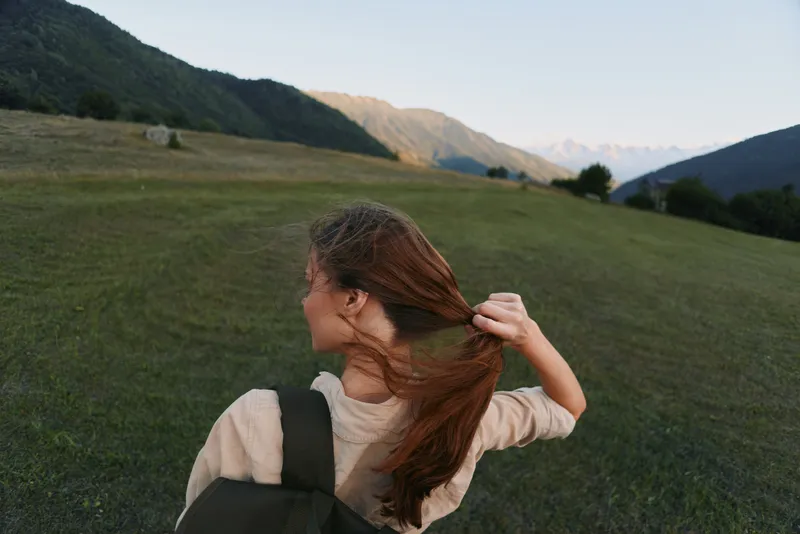 Young woman with a backpack stands in a expansive green field, pulling her long hair back as distant mountains glow in the late light, conveying freedom