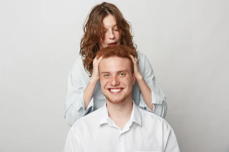 Redhead man with freckles smiles while woman with curly hair massages his head. Casual clothing and light background create friendly and relaxed atmosphere. Concept of friendship and care.