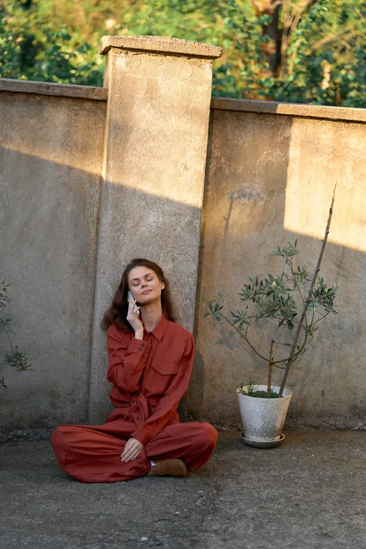 Relaxed woman lifestyle outdoor portrait seated against a stone wall in a red outfit, casual fashion, calm serenity, bright natural light, candid mood, warm ambiance, simple composition