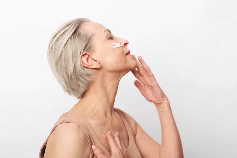Mature woman with short grey hair applies face cream on cheek, showing skincare routine and self-care concept with soft natural lighting on white background.