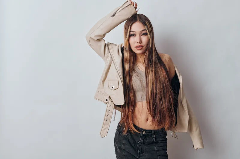 Woman with long hair wearing casual clothes posing against light background. Stylish female model with jacket on shoulder and confident expression in studio photo shoot.