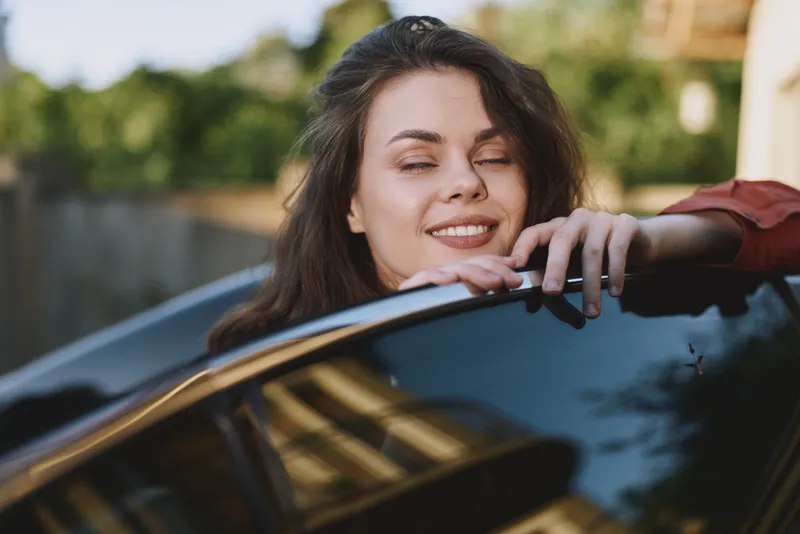 Woman leans on a sleek car, smiling with relaxed confidence, capturing a casual lifestyle moment on a sunny day, exuding happiness, freedom, and a contemporary sense of ease in everyday life