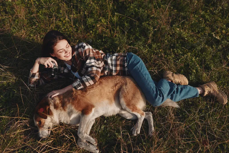 Woman resting beside a relaxed dog in a sunlit grassy field, enjoying quiet outdoor companionship, gentle breeze, and peaceful nature moments shared together.