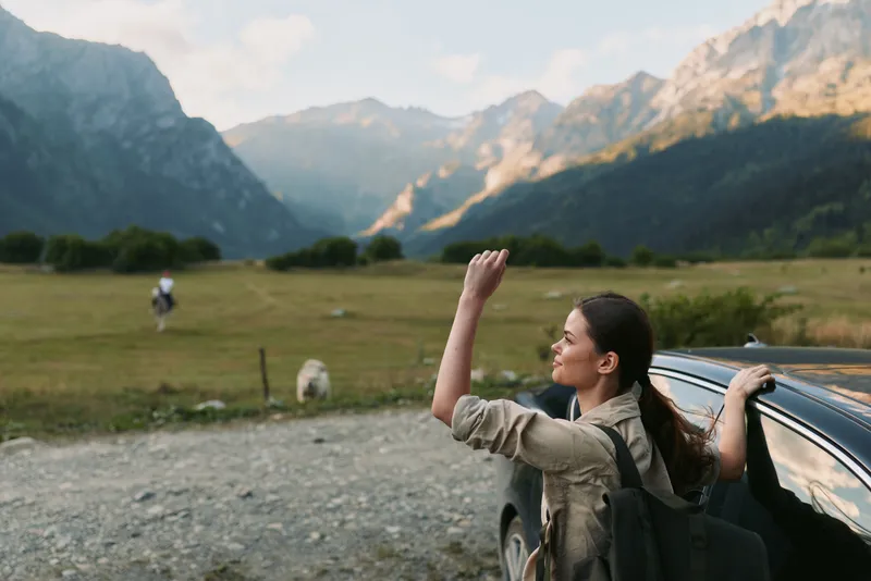 Young woman with long dark hair leans on a car door while enjoying a mountainous landscape during a hiking trip in nature.