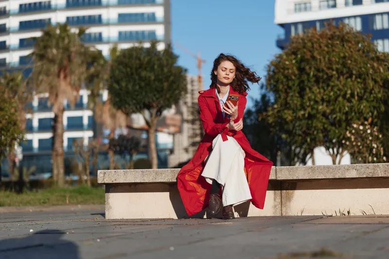 A focused woman sits on a concrete bench outdoors, wearing a red coat over a white dress, checking a phone as wind moves her hair in an urban park.