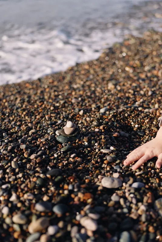 Beach scene with a hand near a small stone stack on a pebble shoreline, calm sea in the background, highlighting balance, focus, and mindfulness in outdoor travel.