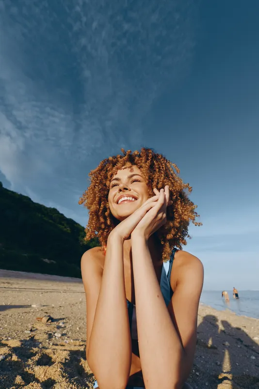 Happy smiling woman with curly hair enjoys sunny day on sandy beach near sea, wearing swimsuit, feeling relaxed and peaceful under blue sky with clouds.