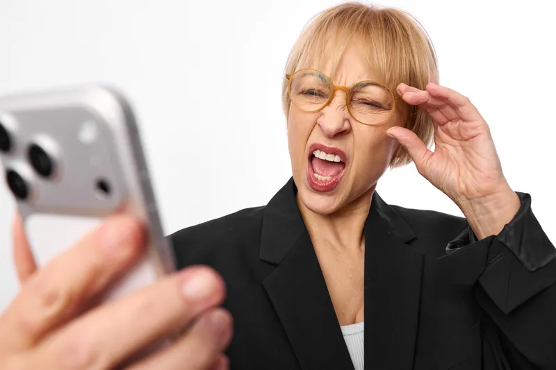 Surprised senior businesswoman with glasses raising a hand to her eyewear while holding a smartphone, shouting in office style, white background, closeup portrait