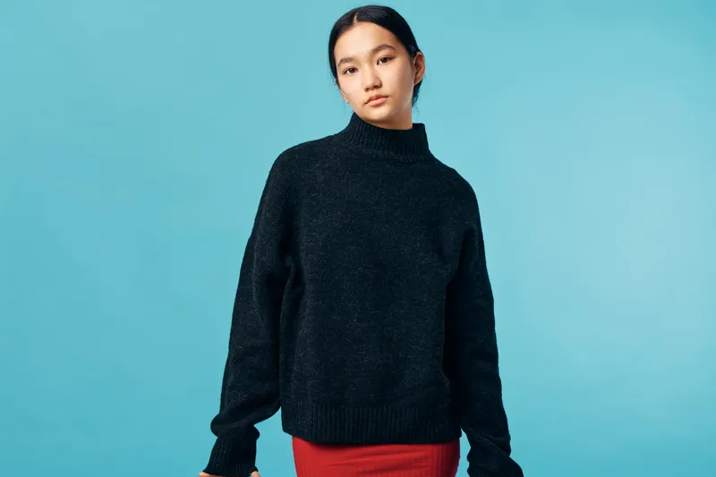 Young woman with black hair wearing a dark turtleneck sweater and red skirt standing against blue background in studio. Casual fashion portrait of confident female model with natural makeup and hair.