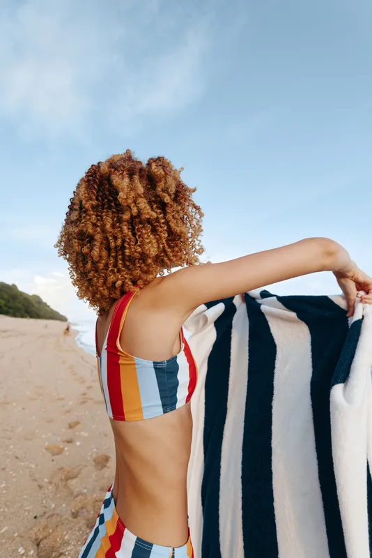 Woman with curly hair wearing colorful striped swimsuit holding striped towel on sandy beach under clear sky. Summer vacation, relaxation, and outdoor leisure concept.