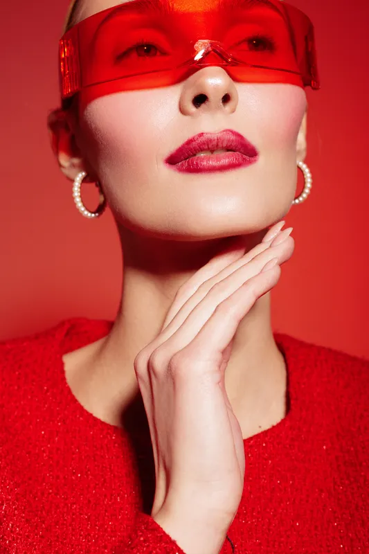 Close-up portrait of a stylish woman wearing red sunglasses and red lipstick with pearl earrings. Fashion concept with vibrant red tones and elegant makeup on a red background.