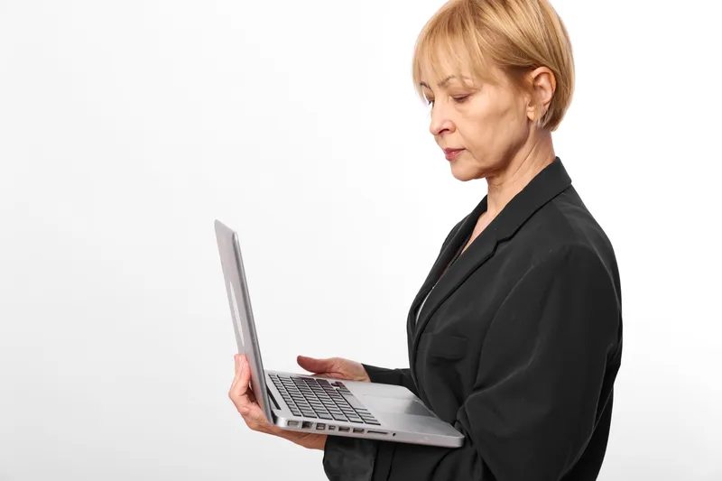 Business woman holding laptop, focused expression, looking at screen, black suit, standing on plain white background, professional office worker preparing report, copy space